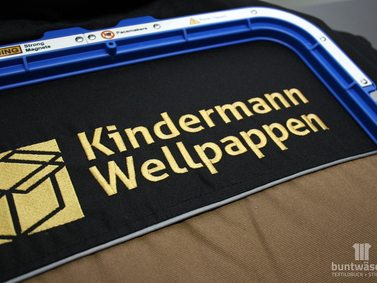 Goldfarbener Schriftzug Kindermann Wellpappen, gestickt auf schwarze Arbeitskleidung
